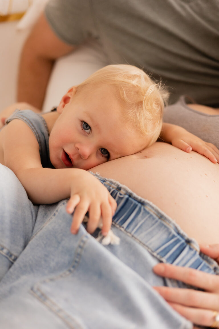 Toddler boy hugging pregnant mother's belly looking at the photographer with blue eyes.