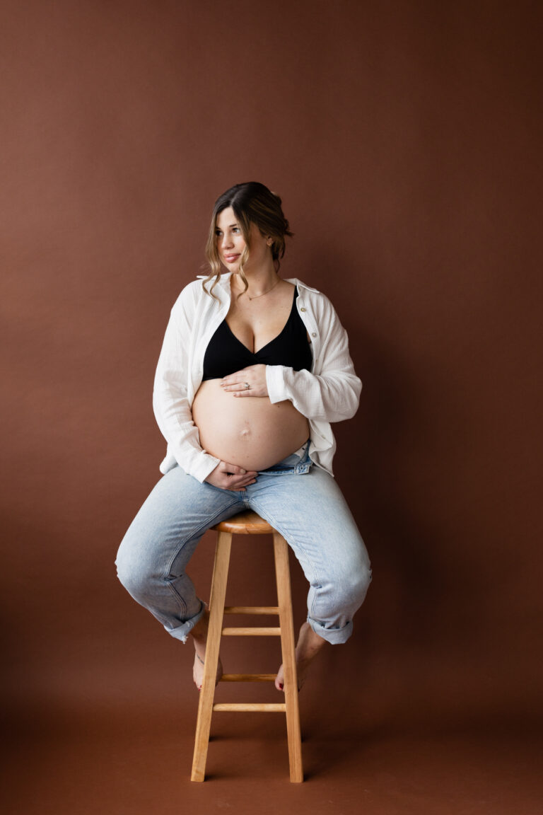 pregnant woman on stool in photography studio holding her stomach for maternity photos