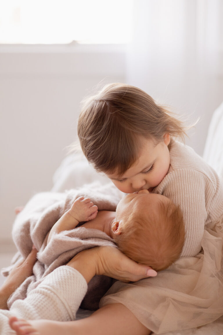 Toddler girl kissing newborn baby brother while holding him on a couch in a photography studio for a newborn session