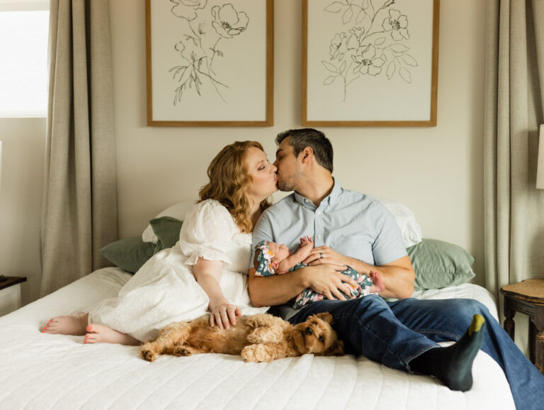 Wife and husband kiss on their bed while holding their newborn baby girl and petting their dog in their home in Monroe, WA for their photography session