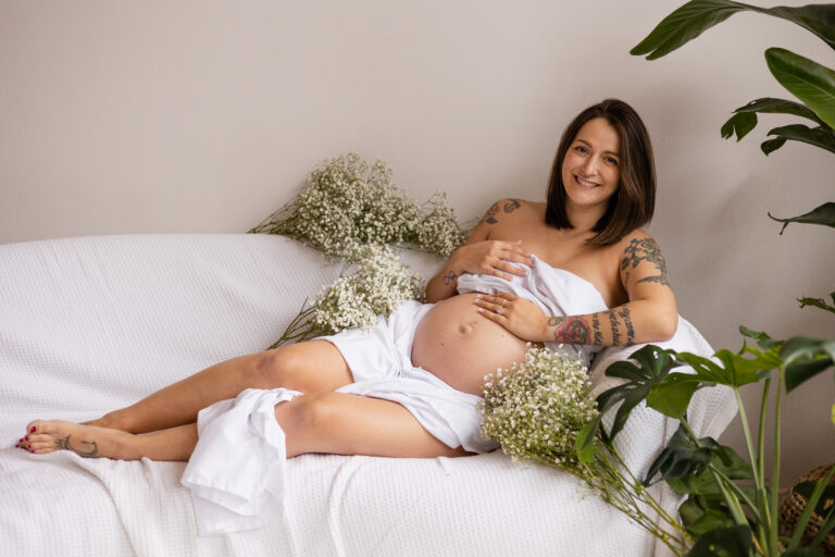 Intimate maternity session of a pregnant mother on a couch wearing only a white sheet surrounded by flowers