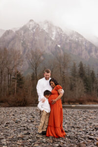 Family with young son and pregnant mom hugging in North Bend by the river with Mt. Si behind them for maternity photoshoot