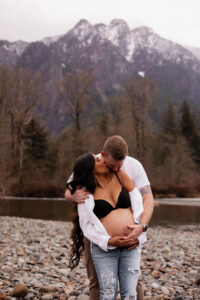 Mom and dad hug and kiss each other while holding moms pregnant belly by the river with Mt. Si behind them during pregnancy photoshoot
