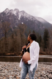 Pregnant mom holds belly and looks back at Mt. Si behind her by the river during maternity photo session in north Bend, WA