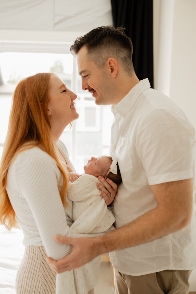 new mom and dad smiling at each other holding their newborn baby in between them during Seattle in home photo session