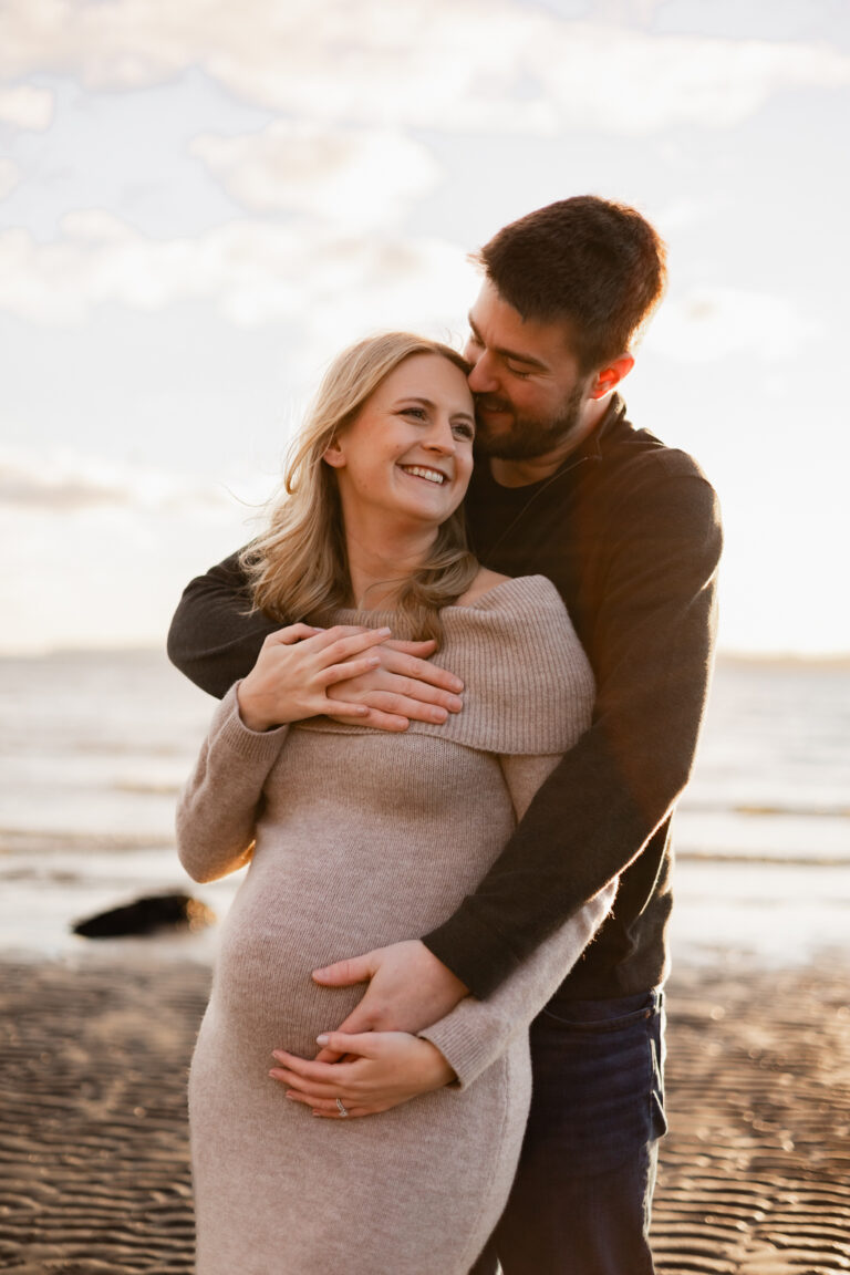 Expecting parents hug and smile at each other during sunset at Discovery Park beach in Seattle for maternity session