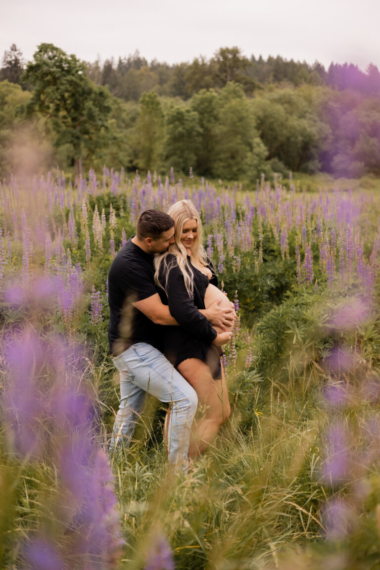 Husband hugs pregnant wide from behind in a field full of lupines in Redmond WA for maternity session