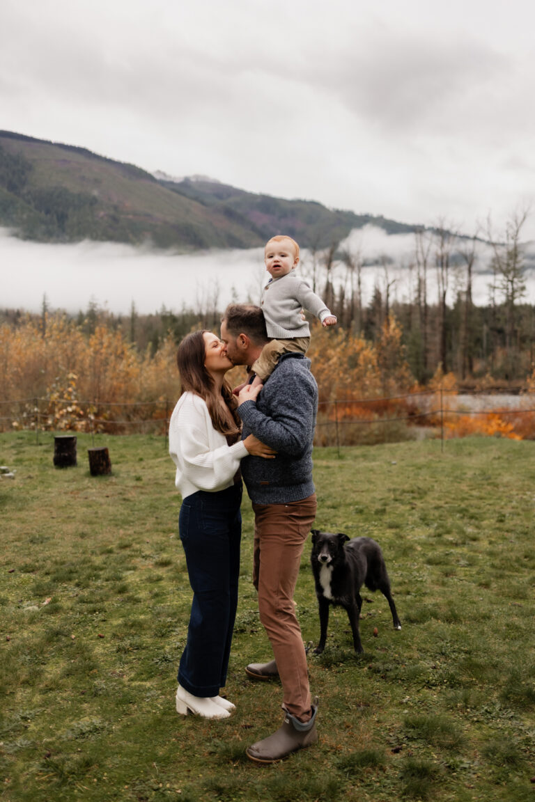 Mom and dad kiss with toddler on the dad's shouldners and their dog by their side near Skykomish river off hwy 2 in the PNW for family photoshoot