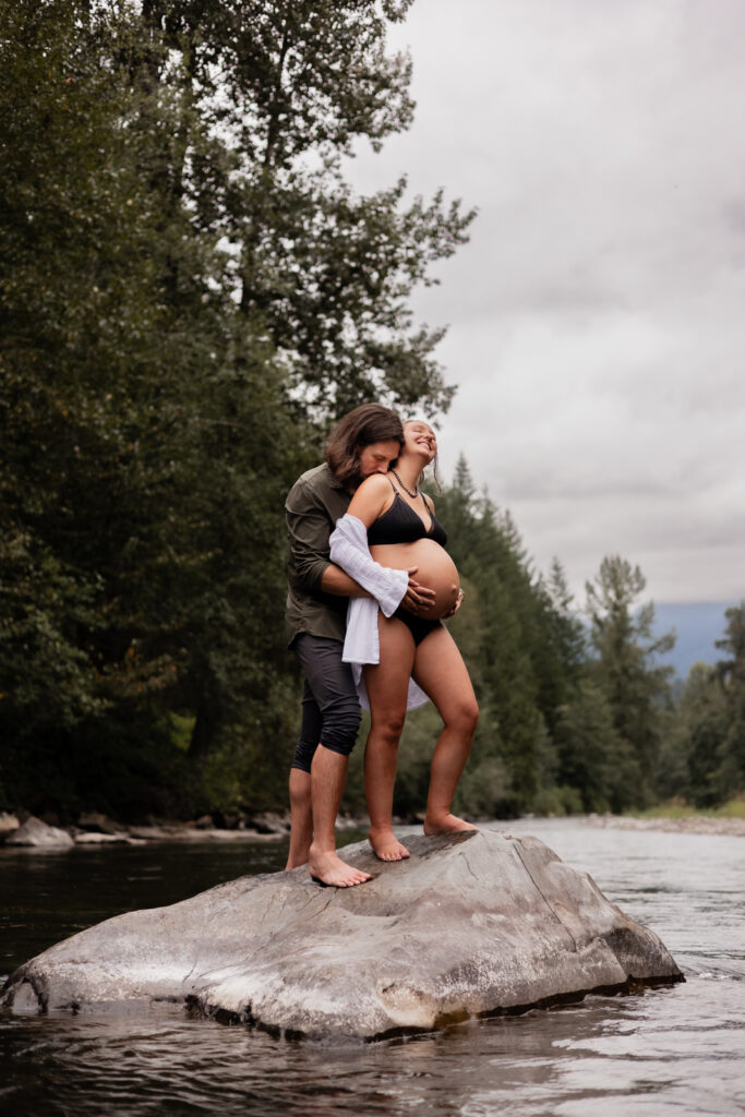 Expecting couple hugs romantically on a rock in the river in North Bend, WA for maternity photography shoot